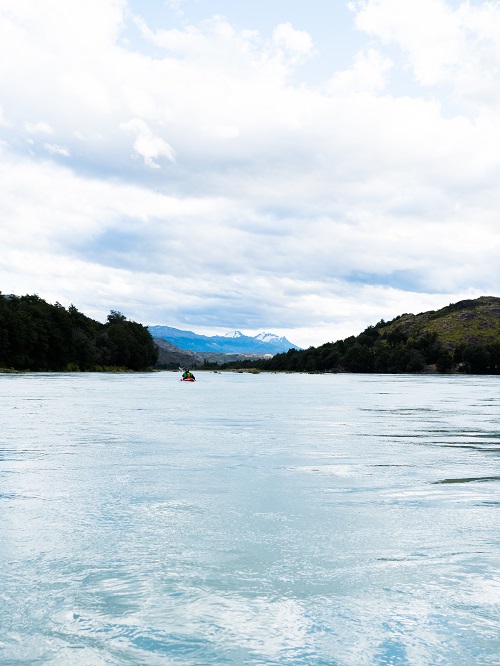 Kayaker in the middle of a calm river with some mountain peaks in the background 