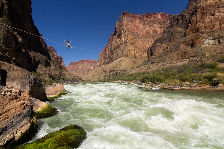 Person kneeling on a slackline above the river with his arms raised in the air with canyon walls all around 