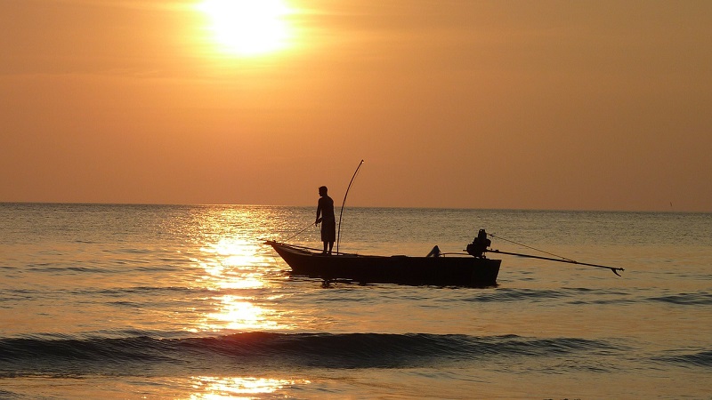 Two people fishing off a boat in the ocean while the sun sets. 