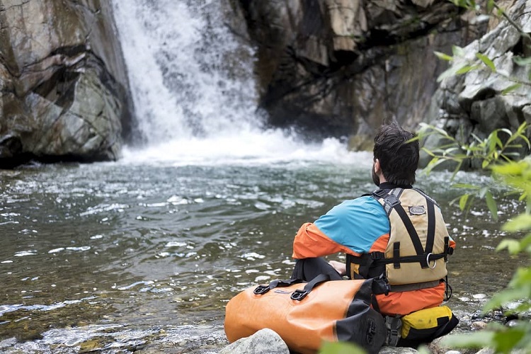 Man sitting on a rock with his PDF on and a Watershed Drybag next to him watching a waterfall 