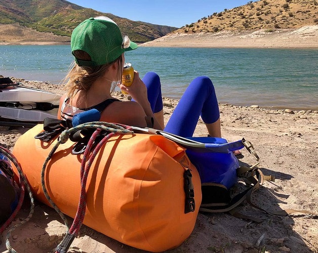 Women leaning against a Watershed Drybag on the shore overlooking the river